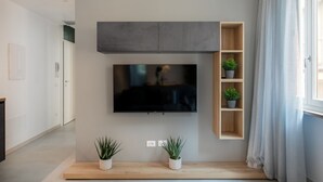 A modern living room with a flat-screen TV mounted on the wall, wooden shelving with plants, and a minimalist design.