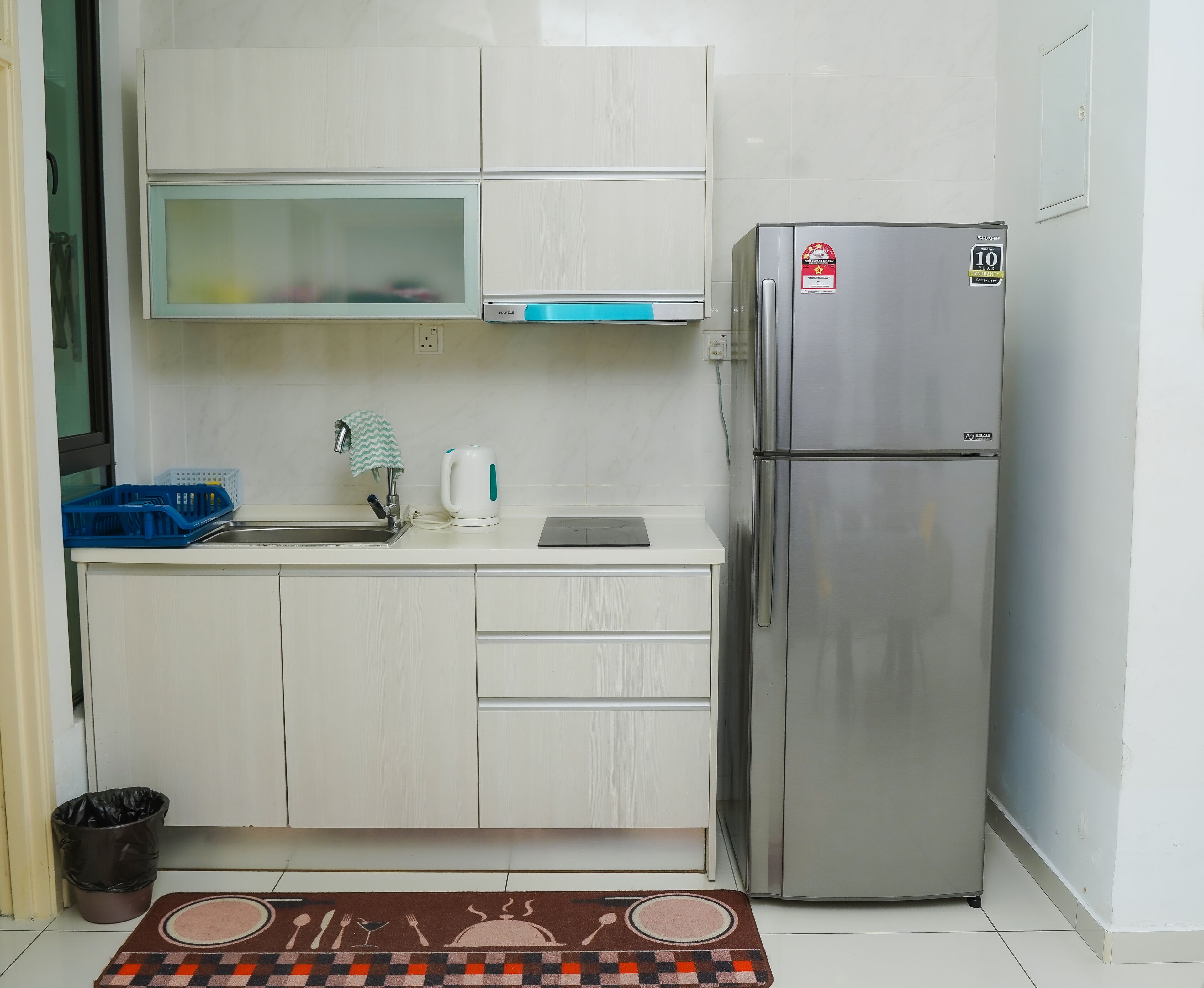 A compact kitchen with a stainless steel refrigerator, a microwave, and a kettle on the countertop. There is a trash bin and a small rug with a table setting design.