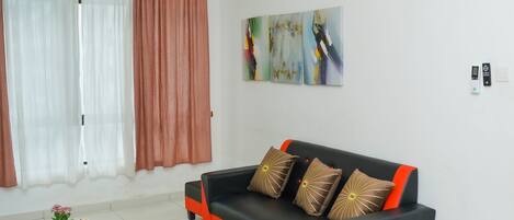 Una sala de estar con sofá, una mesa de centro, un tapete estampado, un cuadro en la pared y una zona de comedor con vajilla y tazas rosas.