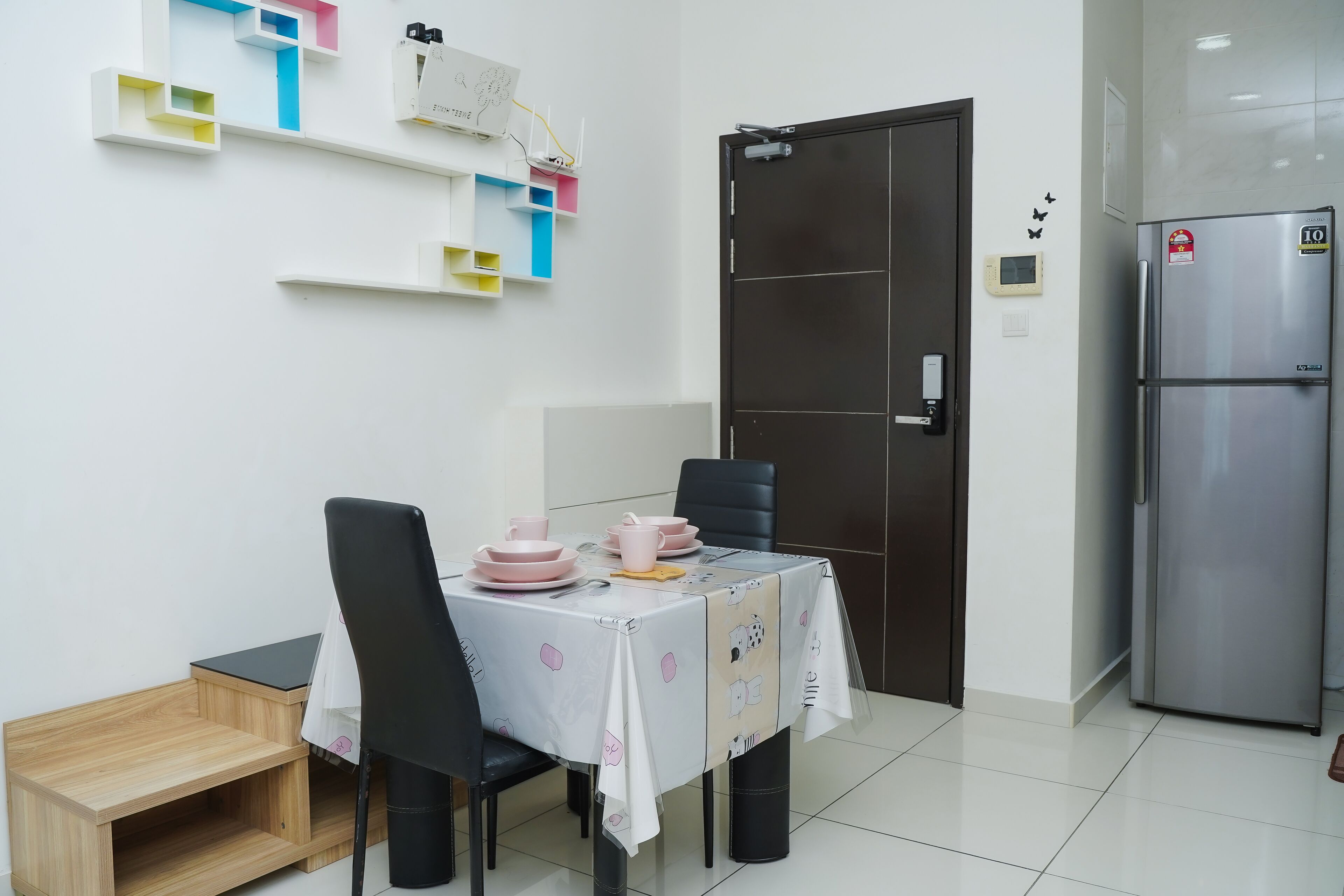 A dining area with a table set for four, chairs, a small wooden side table, a refrigerator, and a door with a lock.