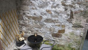Un muro de piedra con celosía metálica, dos sillas, una mesa y un florero con flores.