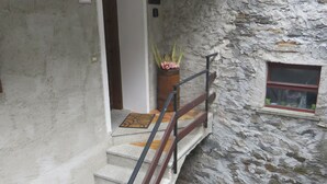 Una escalera con barandilla que lleva a una puerta, una ventana con vistas y un pequeño espacio exterior con sillas y una mesa.
