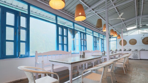 A dining area with white wicker chairs, a long table, and hanging pendant lights. - MANJARI RESORT (Ghansyali)