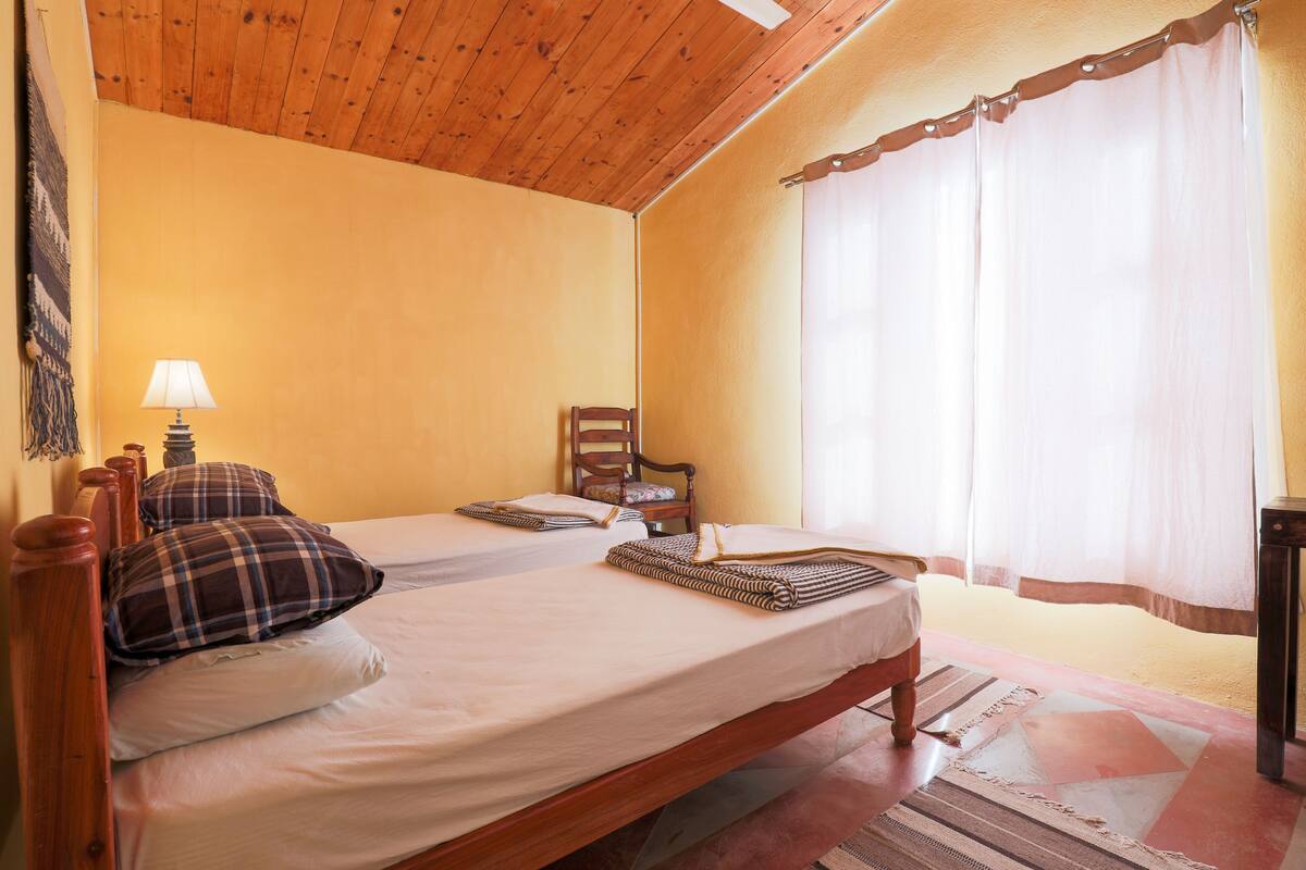 A bedroom with two beds, wooden headboards, a chair, and a window with curtains.