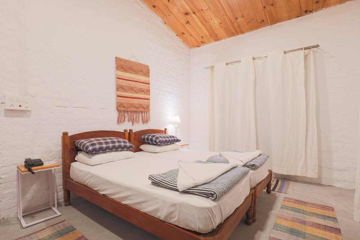 A bedroom with a wooden bed, white walls, a wooden ceiling, a bedside table with a lamp, and a patterned rug.