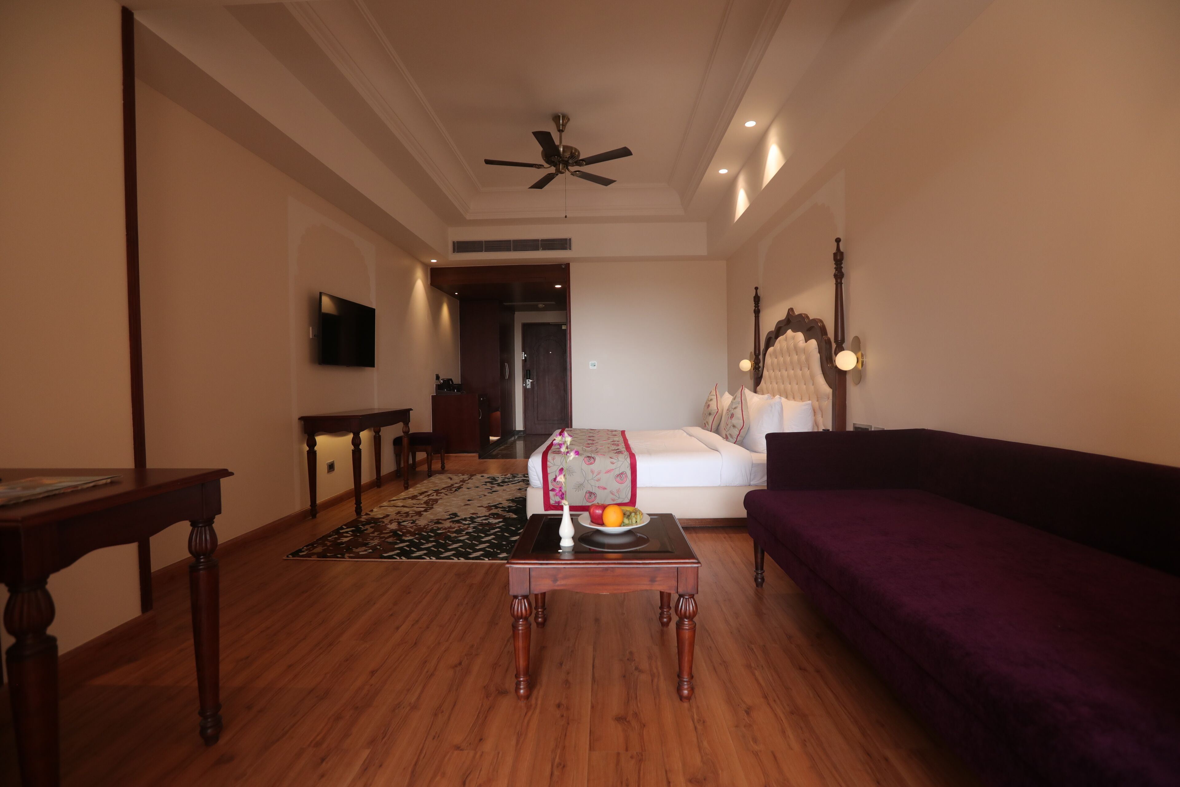 A living room with a wooden floor, a sofa, a coffee table with fruit, a TV mounted on the wall, and a ceiling fan.