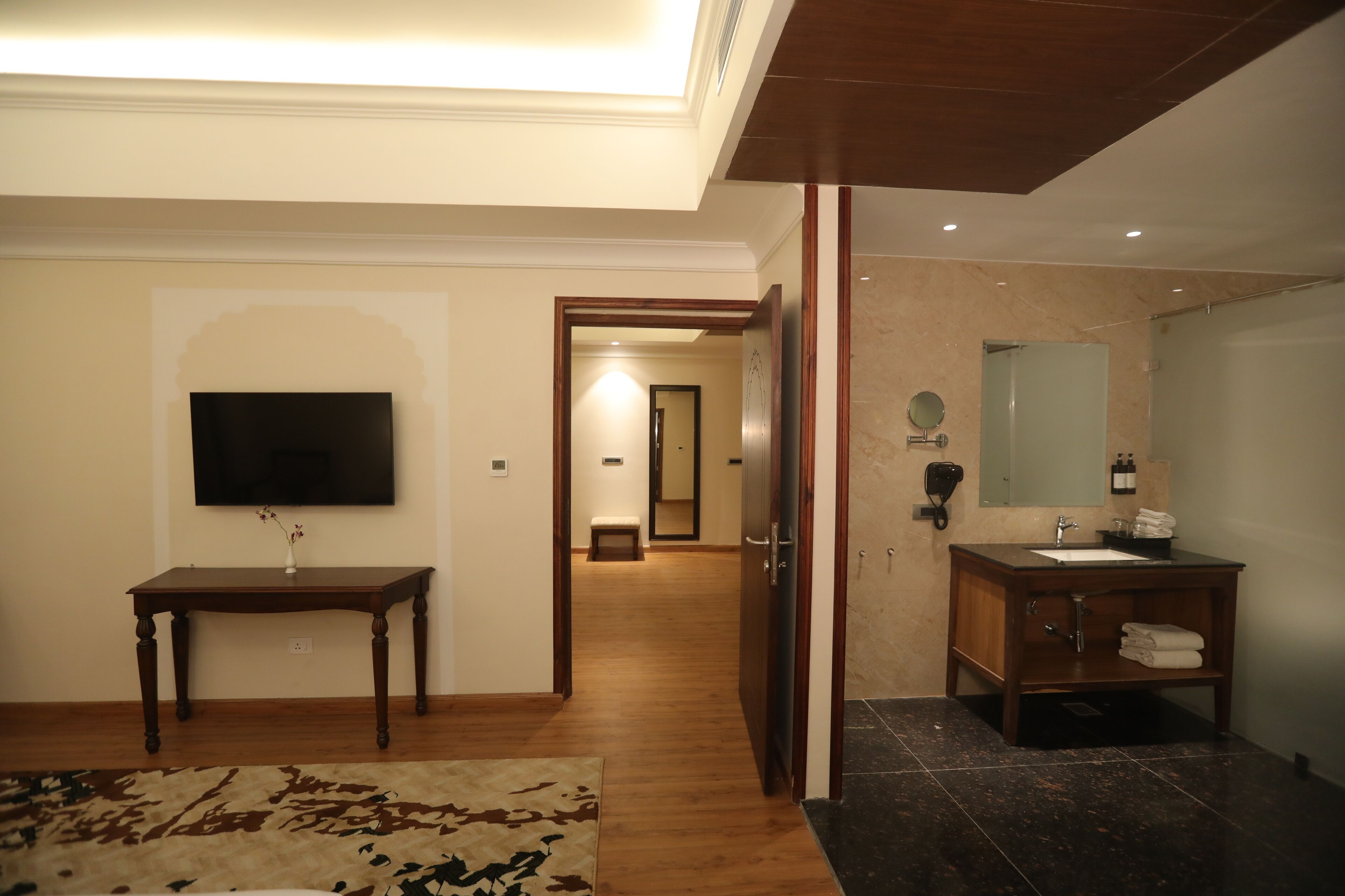 A hotel room with a wooden floor, a flat-screen TV mounted on the wall, a wooden vanity with a mirror, and a small bench in the background.