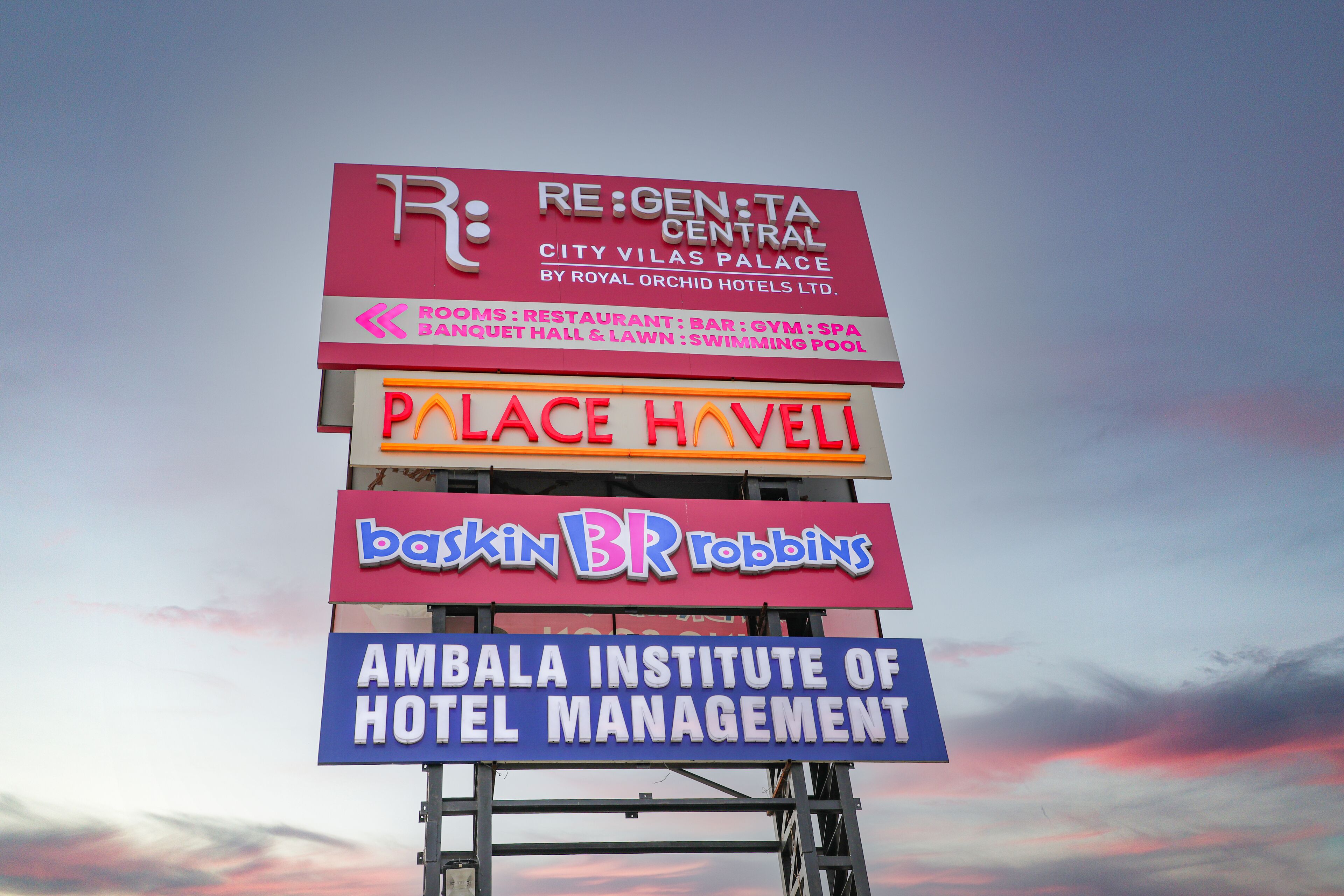 Ein Schild mit mehreren Firmennamen, darunter RE:GEN:TA CENTRAL, Palace Havel, Baskin BR Robbins und das Ambala Institute of Hotel Management.
