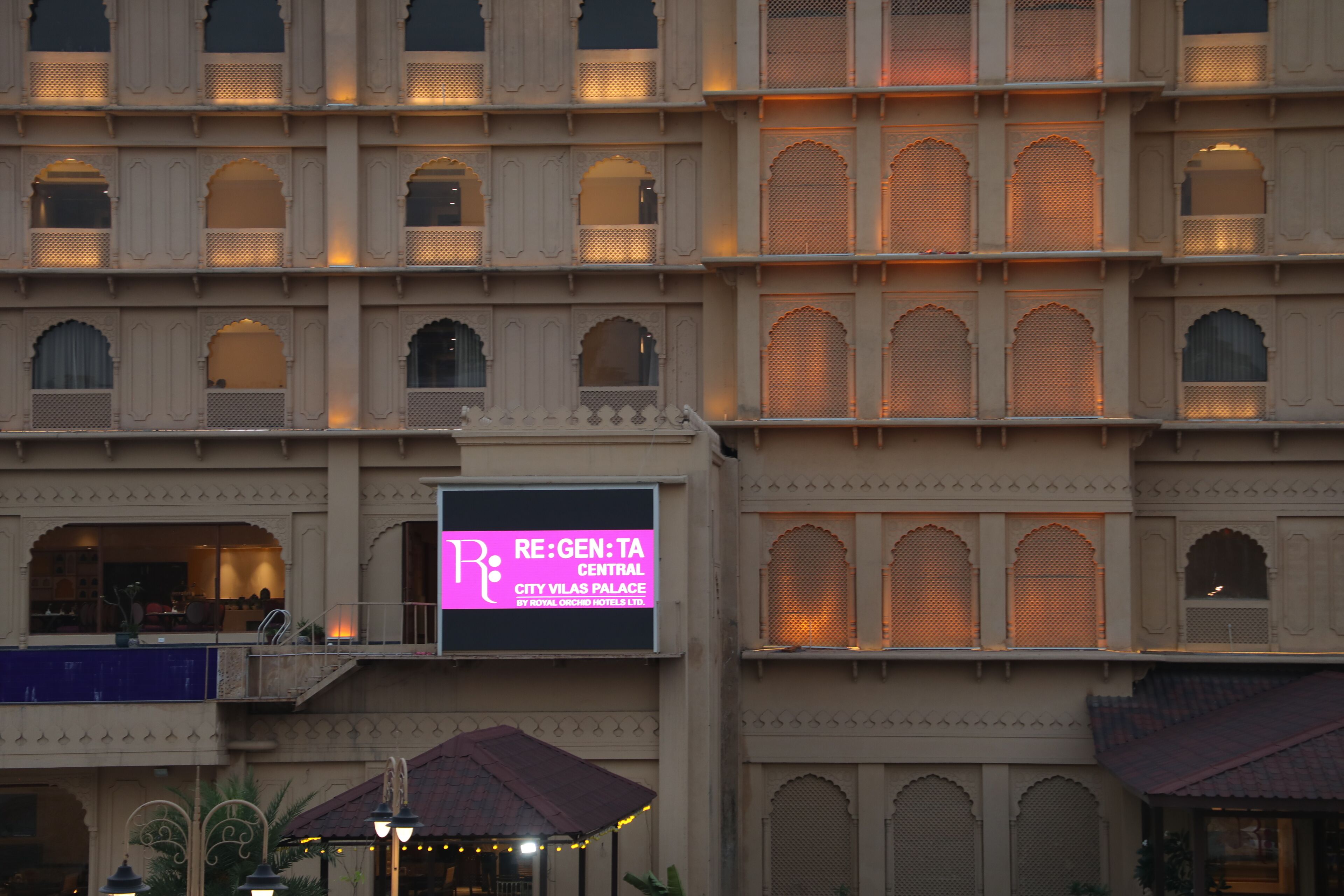 Un immeuble à plusieurs étages doté de fenêtres en arc de cercle et de balcons. Une enseigne indique : « RE:GEN:TA CENTRAL CITY VILAS PALACE BY ROYAL, ORCHID HOTEL LTD. »