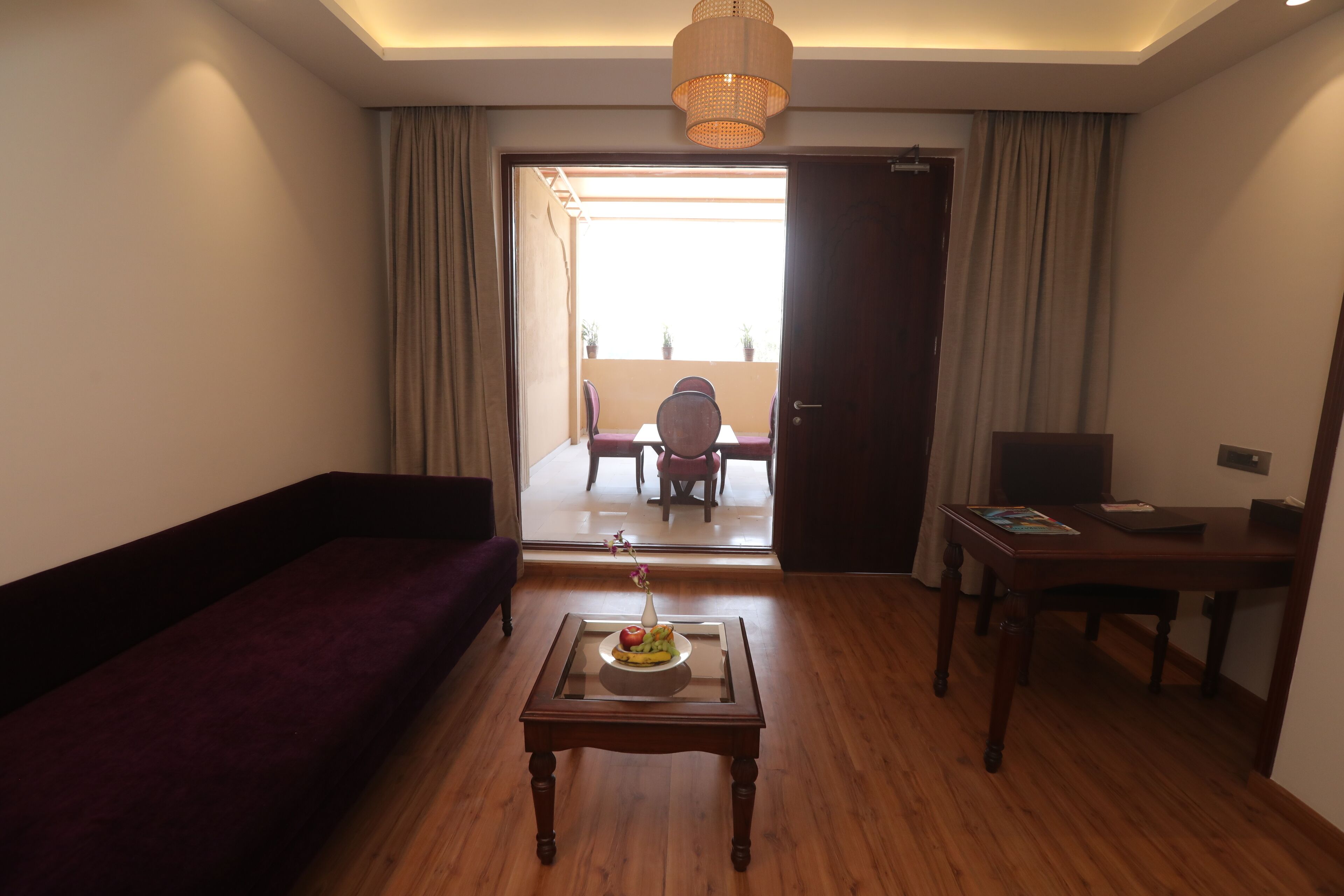 A hotel room with a wooden floor, a purple sofa, a glass-top coffee table with a vase and fruit, a wooden desk with a laptop, and a view of a balcony with a dining table and chairs.