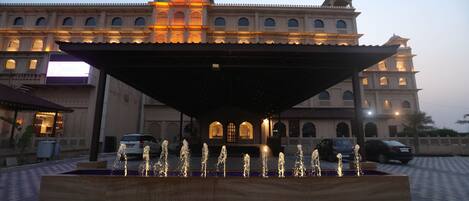 A large building with a fountain in front, a covered area, and parked cars.