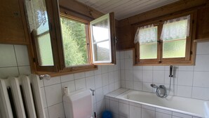 Un baño pequeño con inodoro, lavamanos y bañera. Hay dos ventanas con marcos de madera y cortinas de encaje.