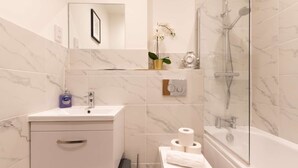 A bathroom with a marble-tiled wall, a white sink with a mirror, a shower with a glass enclosure, and a toilet with toilet paper. - Comfty 1 Bedroom Apartment (Southampton)