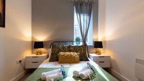 A hotel room with a bed, two bedside tables with lamps, a window with curtains, and a view of a building outside. - Comfty 1 Bedroom Apartment (Southampton)