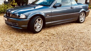 Um BMW conversível azul escuro estacionado sobre cascalho em frente a uma casa com varanda.