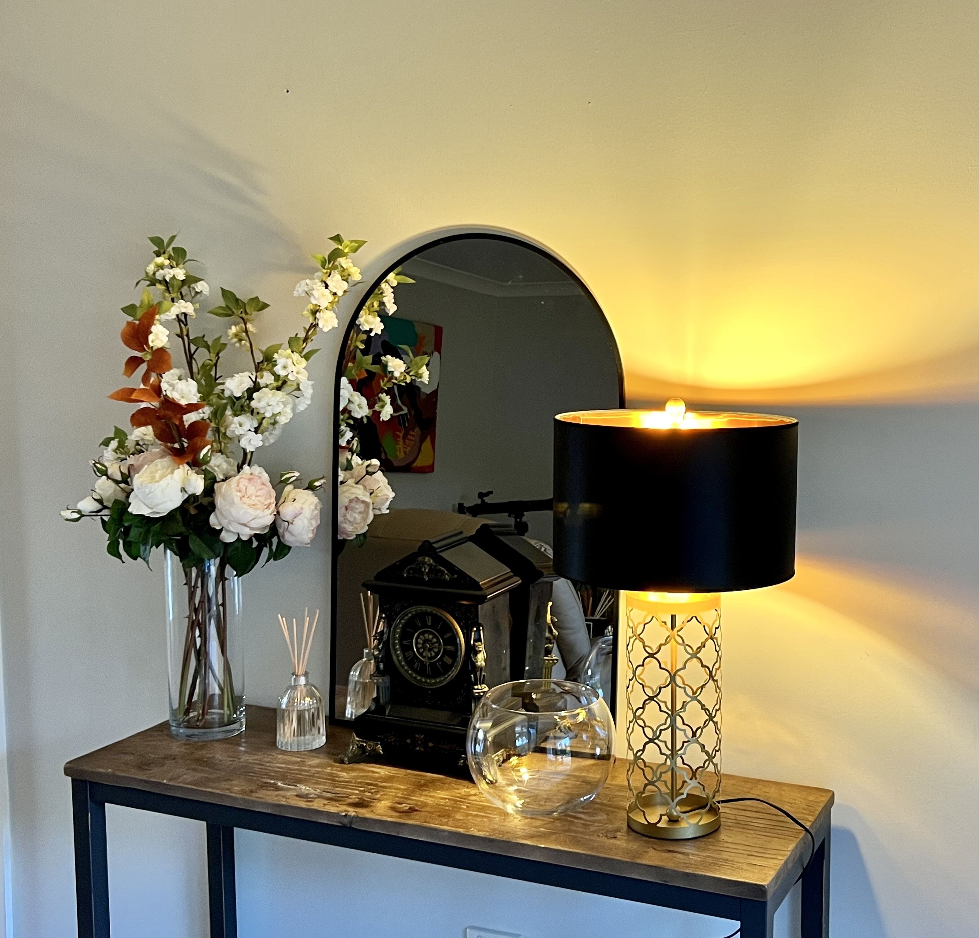 A wooden console table with a black lamp, a vintage camera, a mirror, and a vase with flowers.