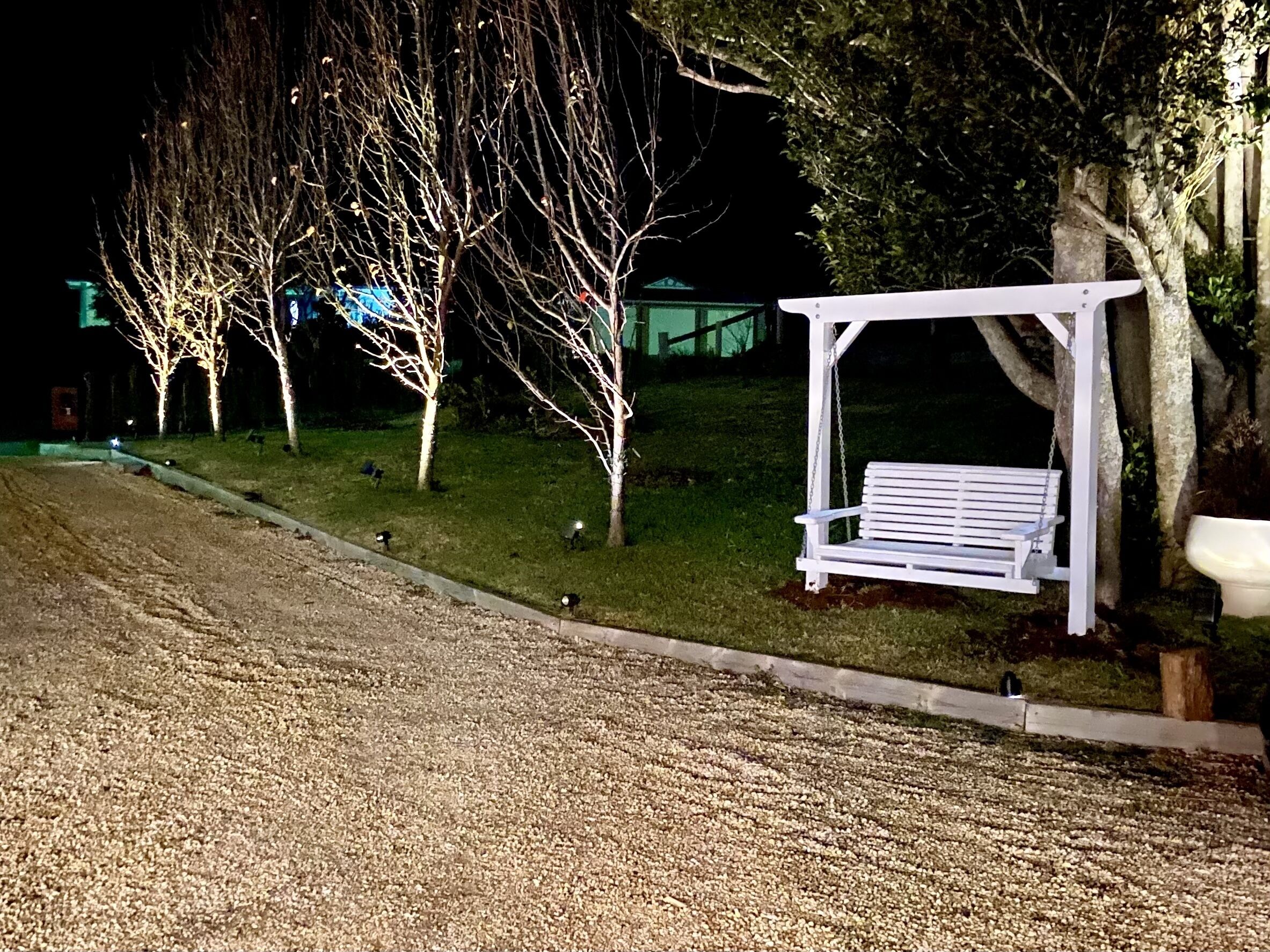 A white wooden swing set under a tree, a gravel path, and leafless trees in the background.