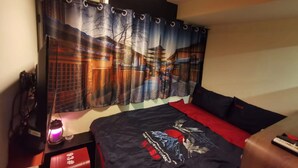 A room with a bed, a red side table, a lamp, and a curtain with a cityscape design. - KMB hostel (Kowloon)
