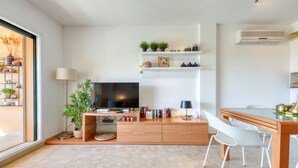 A modern living room with a flat-screen TV, wooden furniture, and a dining area with white chairs.