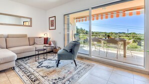 A modern living room with a sofa, a coffee table, and a chair. There is a balcony with a table and chairs, and a view of the countryside.