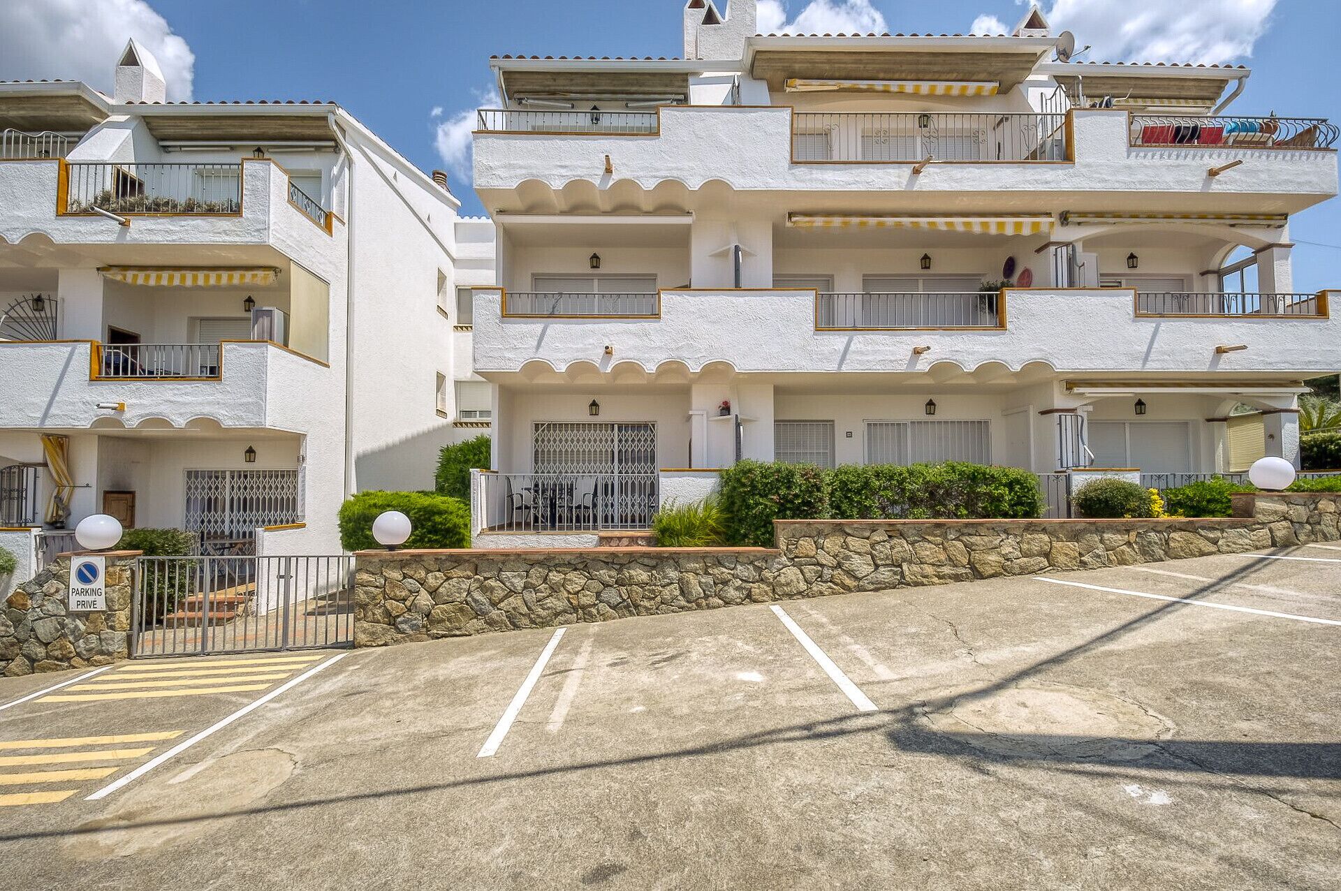 A multi-story residential building with balconies, a stone wall, and a parking area.