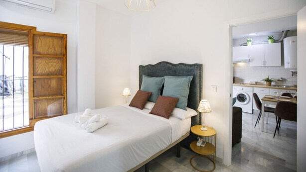 A modern bedroom with a large bed, bedside lamps, and a view of the kitchen. - Rentalsevilla Escapada en San Luis (Sevilla)