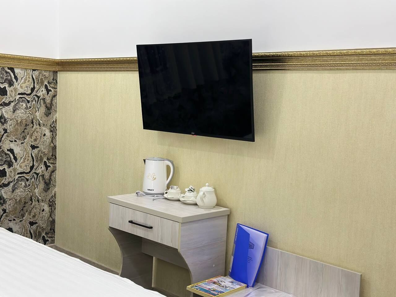 A hotel room with a flat-screen TV mounted on the wall, a small bedside table with a kettle and teapots, and a bed with a white comforter.