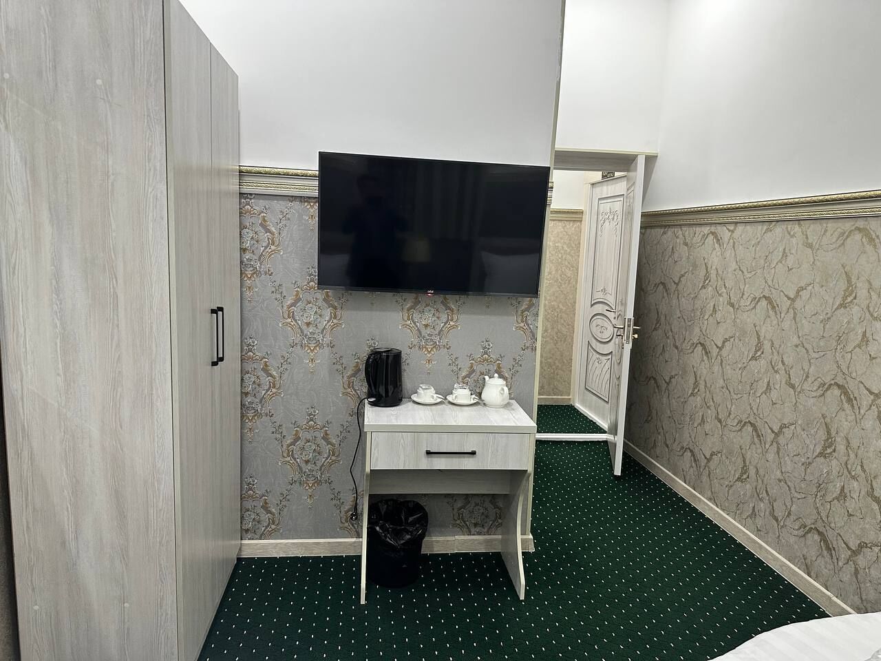 A hotel room with a wardrobe, a desk with a television, a small trash bin, and patterned wallpaper.