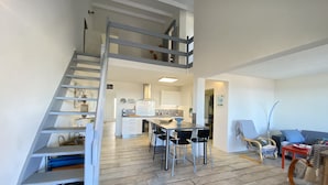 A modern interior with a staircase leading to a loft area, a dining area with a kitchen in the background, and a seating area with a sofa and chairs. - Seafront house (Fleury)