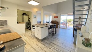 A modern kitchen with a central island, a dining area with bar stools, and a staircase leading to a balcony. - Seafront house (Fleury)