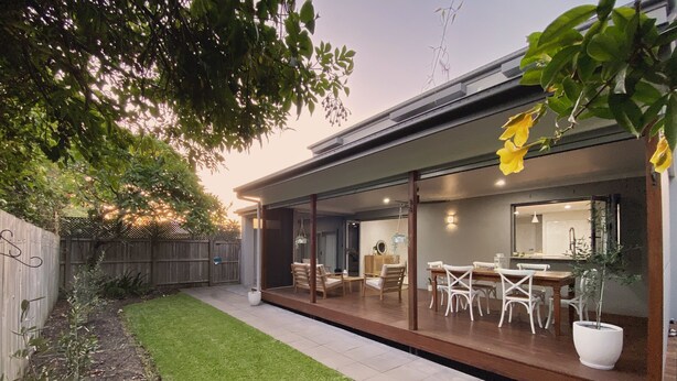 Une maison moderne dotée d’un patio couvert, d’un coin repas et d’une cuisine visible à travers de grandes portes vitrées.