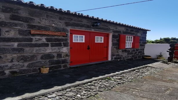 A stone building with a red door and window shutters, a stone pathway, and a small garden area. - Oxen Car House 1 Bedroom (Capelo)