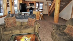 A cozy living room with a fireplace, a sofa, and a coffee table. There is a kitchen in the background with a refrigerator, stove, and microwave. A wooden staircase leads to an upper level. - The Hideaway Hostel near Yellowstone (Island Park)