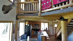 A rustic cabin interior with a wooden staircase, a mounted deer head, a kitchen area, and a dining space with a round table. - The Hideaway Hostel near Yellowstone (Island Park)