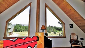A room with a sloped wooden ceiling, two large windows, a red patterned rug, a lamp, and a wooden chair. - The Hideaway Hostel near Yellowstone (Island Park)