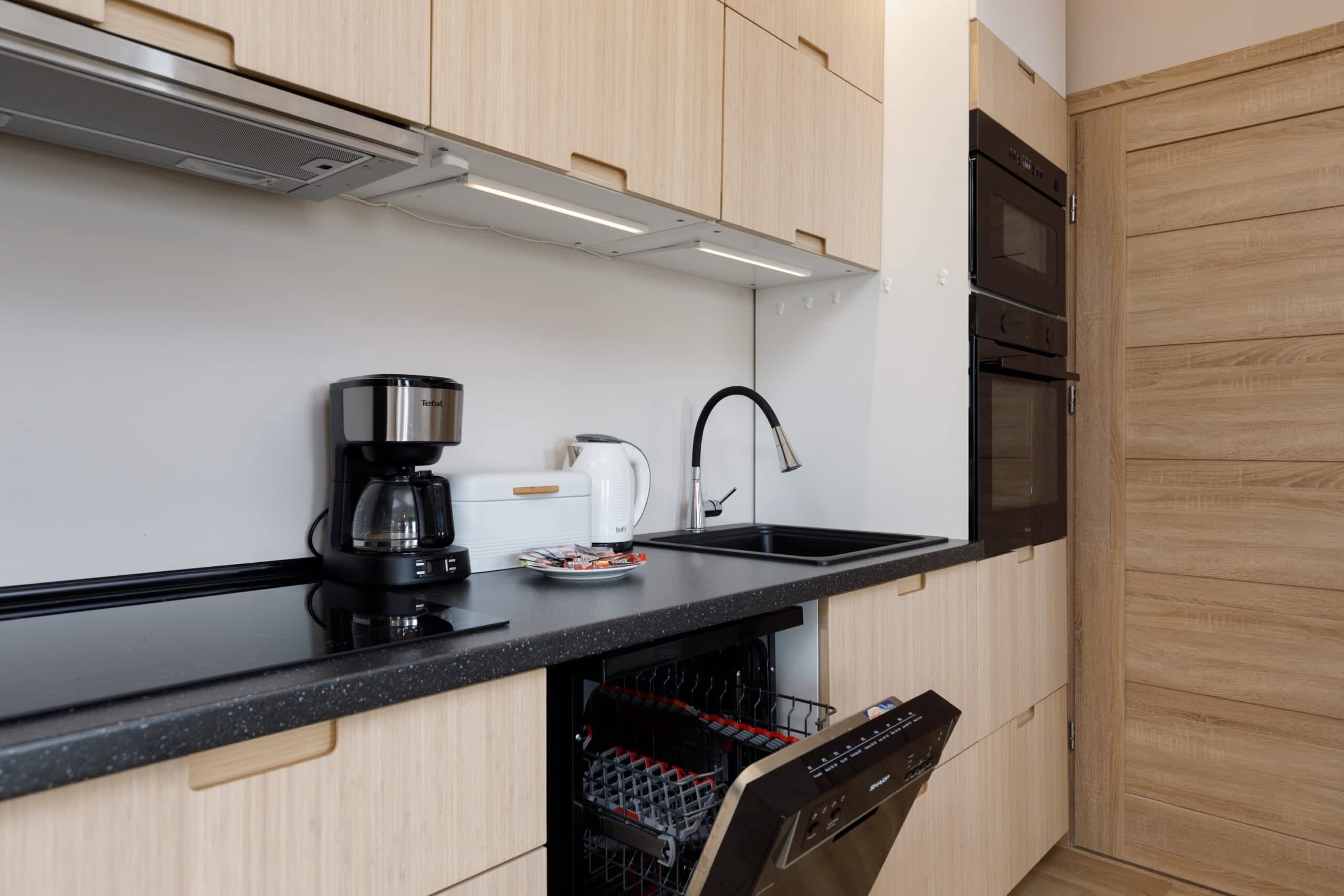 A modern kitchen with wooden cabinets, a black countertop, a coffee maker, a kettle, a dishwasher, and a built-in oven.