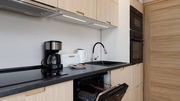A modern kitchen with wooden cabinets, a black countertop, a coffee maker, a kettle, a dishwasher, and a built-in oven.
