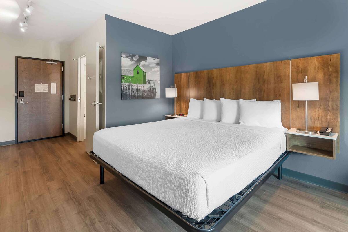 Quarto de hotel com uma cama grande, cabeceira de madeira, duas mesas de cabeceira com luminárias, um quadro na parede e uma porta à esquerda.