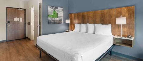 Habitación de hotel con una cama grande, cabecera de madera, dos mesitas de noche con lámparas, un cuadro en la pared y una puerta a la izquierda.