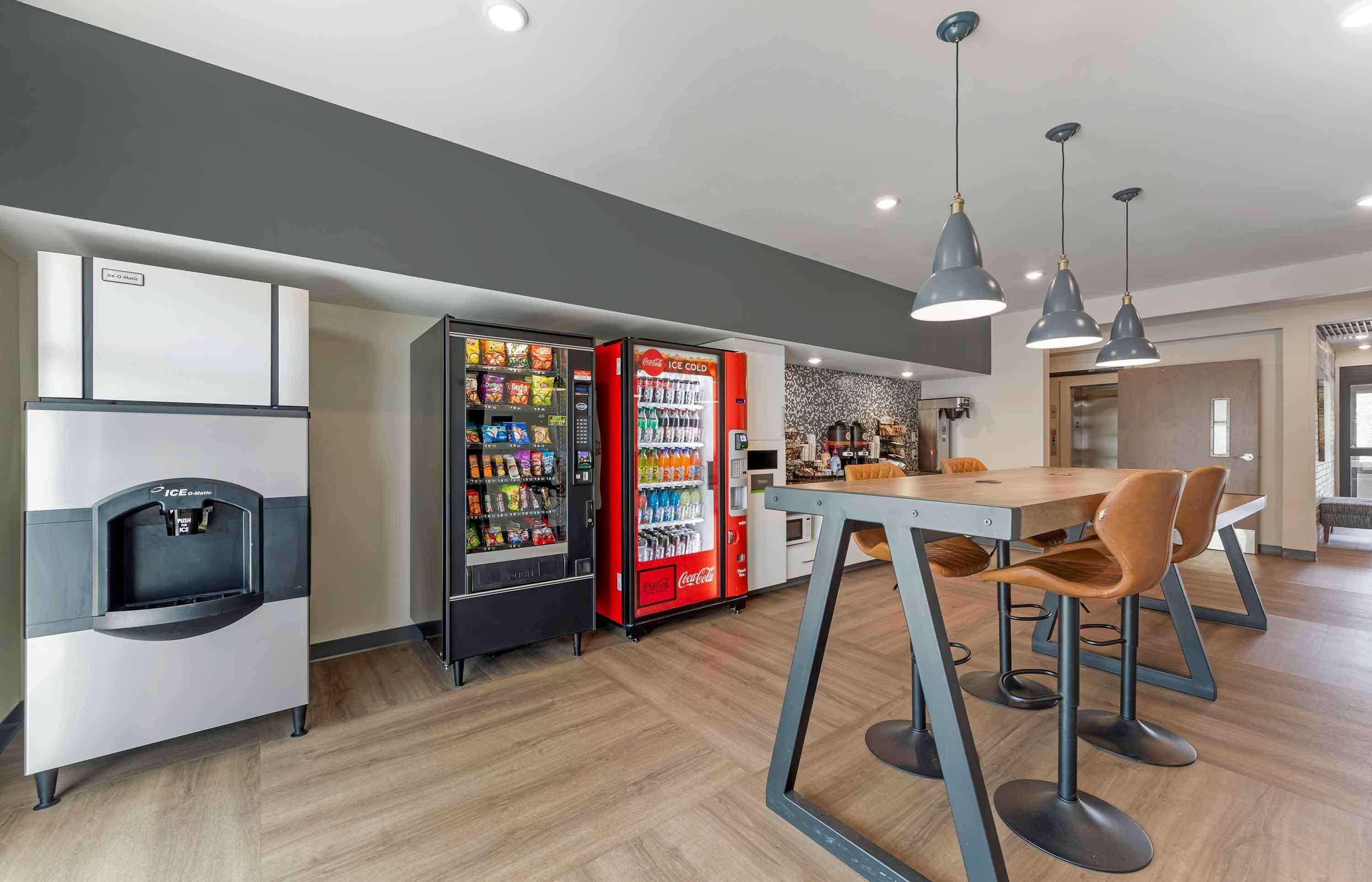 Una cocina moderna con un refrigerador, un refrigerador Coca-Cola y un dispensador de agua. Hay una zona de bar con taburetes y luces colgantes.