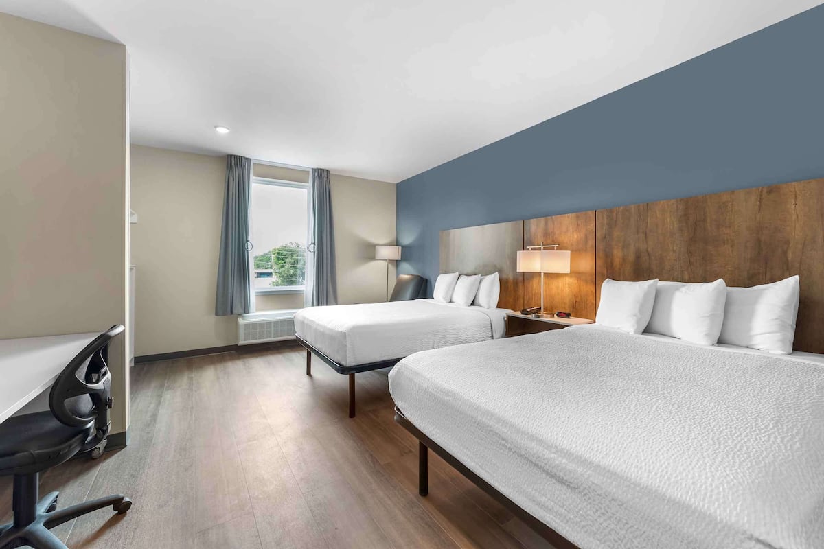 Quarto de hotel com duas camas, uma escrivaninha com cadeira, uma luminária e vista para áreas verdes.