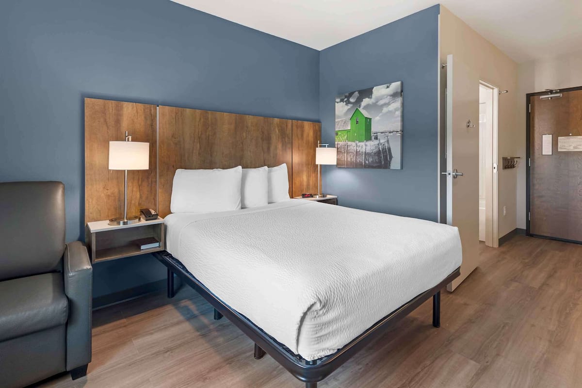 Quarto de hotel com uma cama grande, duas mesas de cabeceira com abajures, uma cadeira e um quadro na parede.