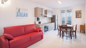 A modern apartment with a red sofa, a dining table with chairs, a kitchen with white cabinets, and a large window offering a view. - Villa Malù - APT Amalia - Pool and lake view (Torri del Benaco VR, Italia)
