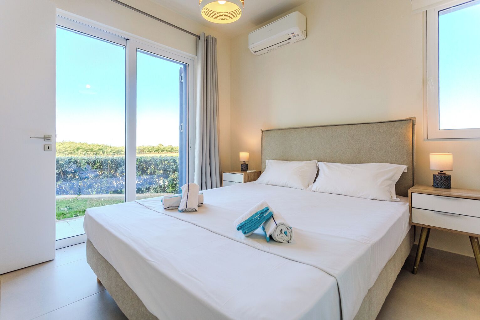 A hotel room with a large bed, a bedside table, a lamp, an air conditioning unit, and a view of a garden through a sliding door.