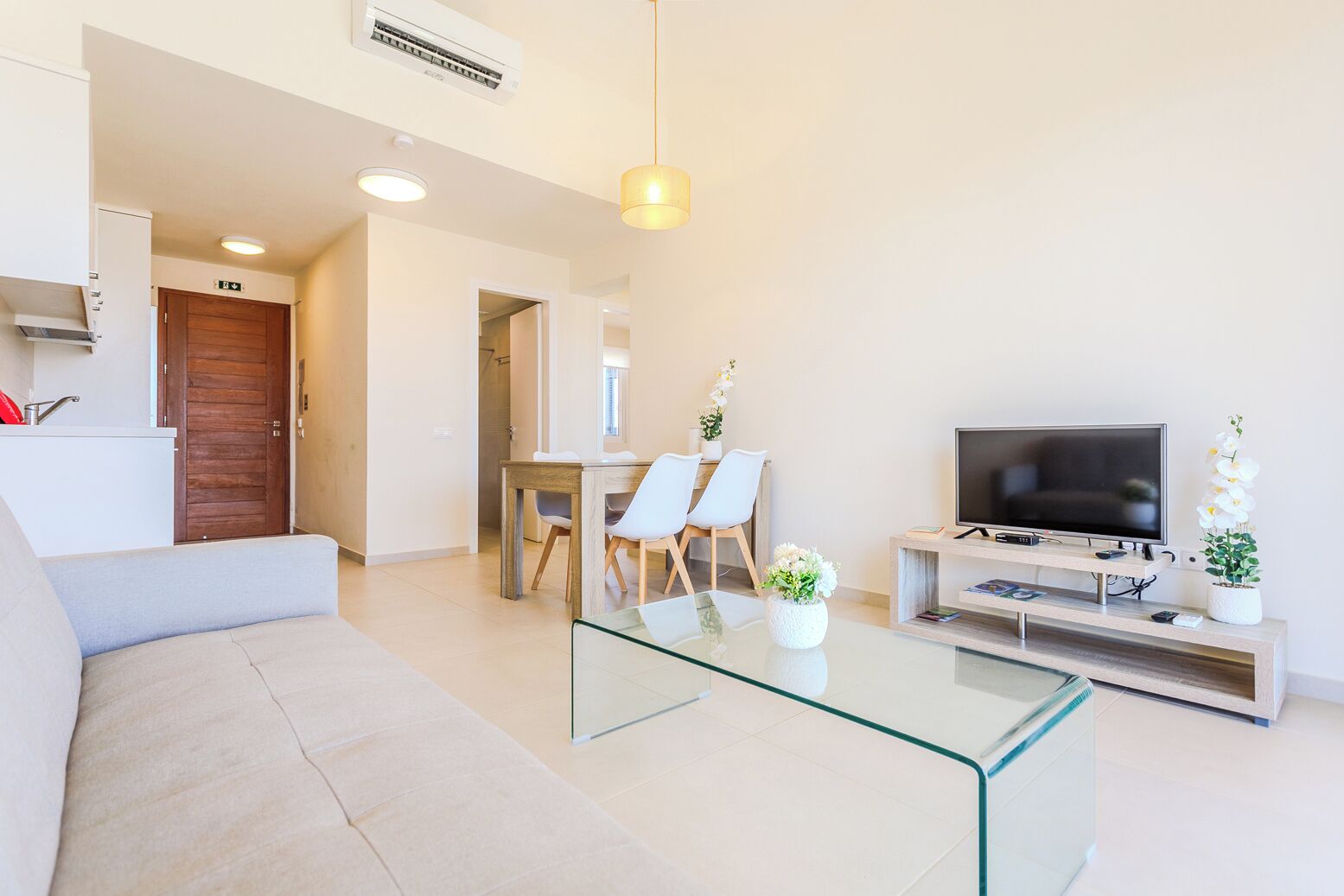Een moderne woonkamer met een glazen salontafel, een bank, een eethoek met stoelen, een televisie op een houten standaard en een airconditioner aan de muur.