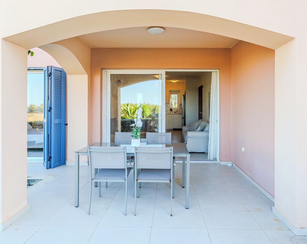 Una terrazza esterna con tavolo e sedie, una porta scorrevole in vetro che dà su un ambiente interno e una porta blu a sinistra.