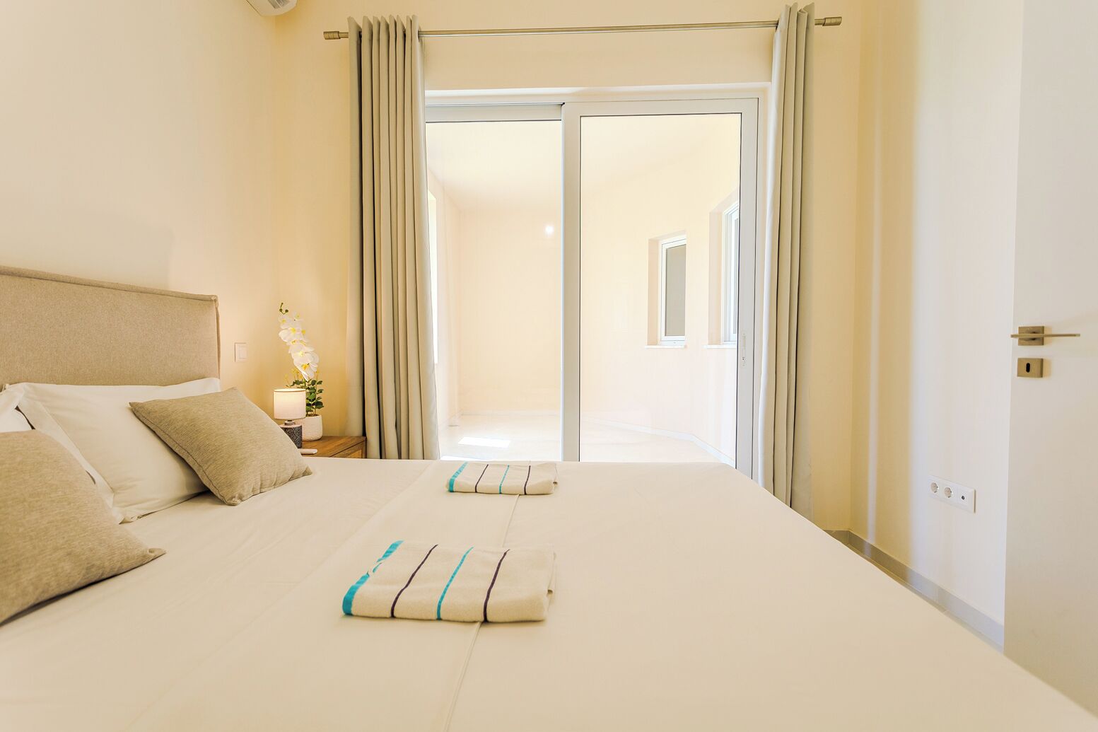 A hotel room with a bed, a nightstand with a lamp, and a view through a door.