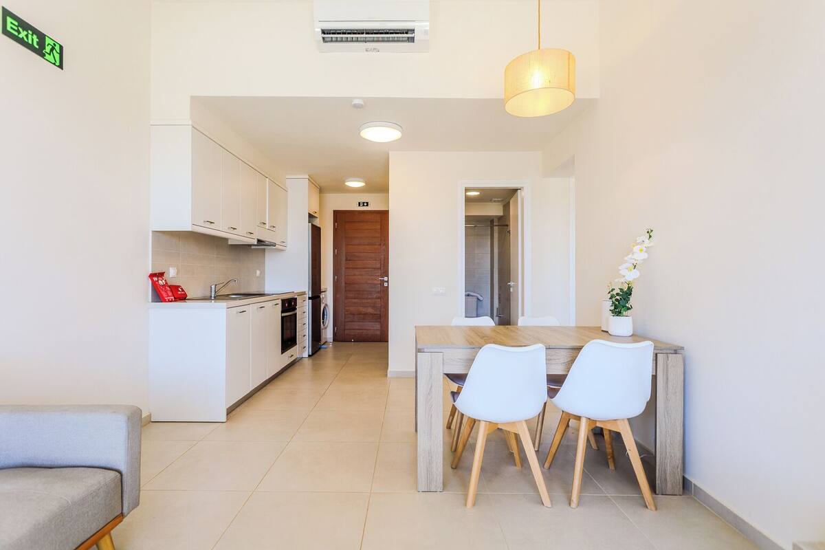 A compact kitchen with white cabinets, a dining area with a wooden table and white chairs, and a visible exit sign.