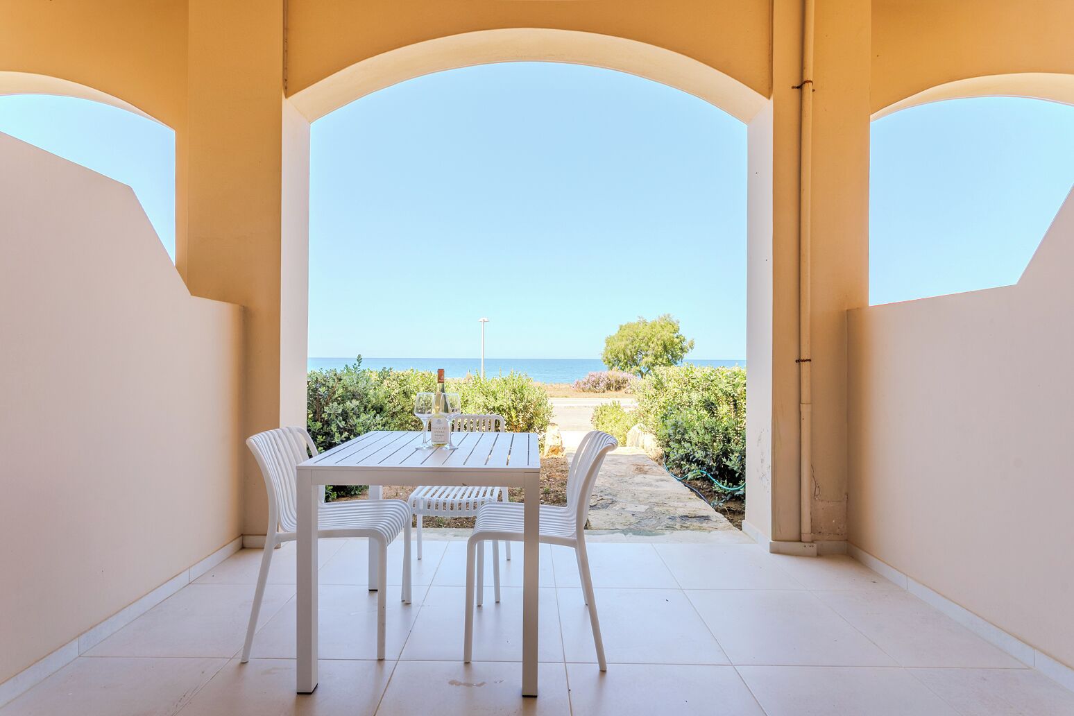 Um alpendre coberto com mesa e cadeiras, vista para o mar e céu claro.