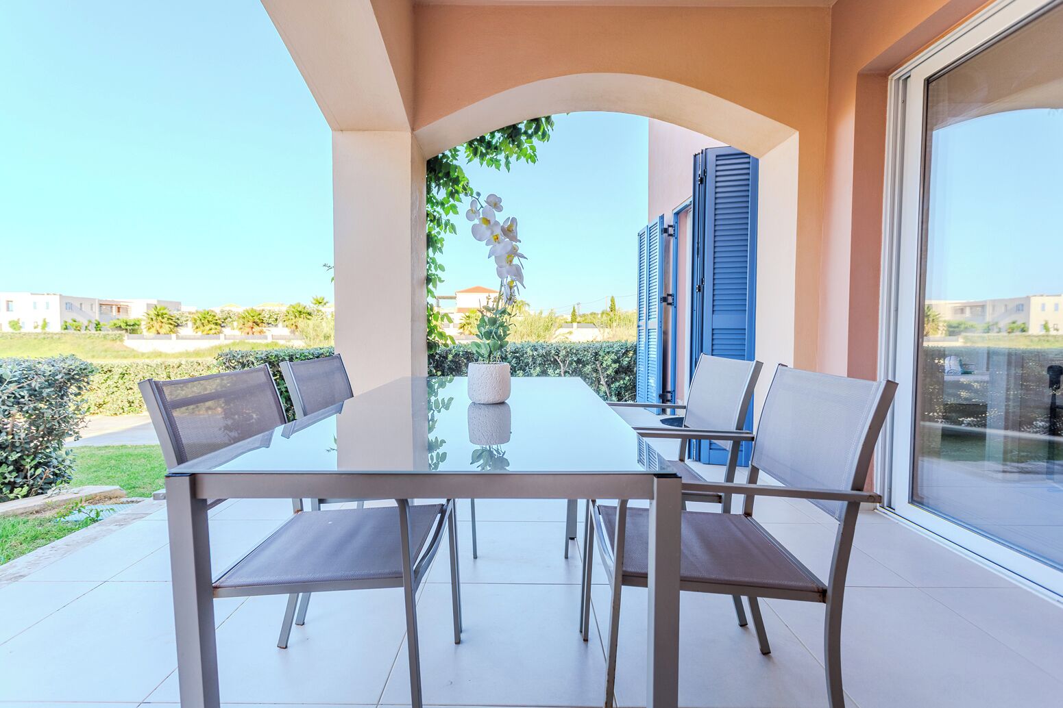 A modern outdoor dining area with a glass table and metal chairs, a potted plant, and a view of a residential area.